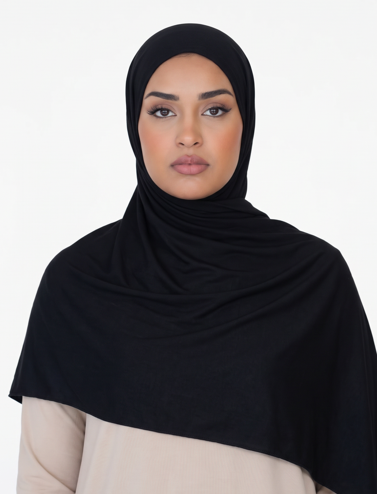 A woman wearing a solid black hijab draped over her shoulders, paired with a light beige top, photographed against a seamless white studio background.