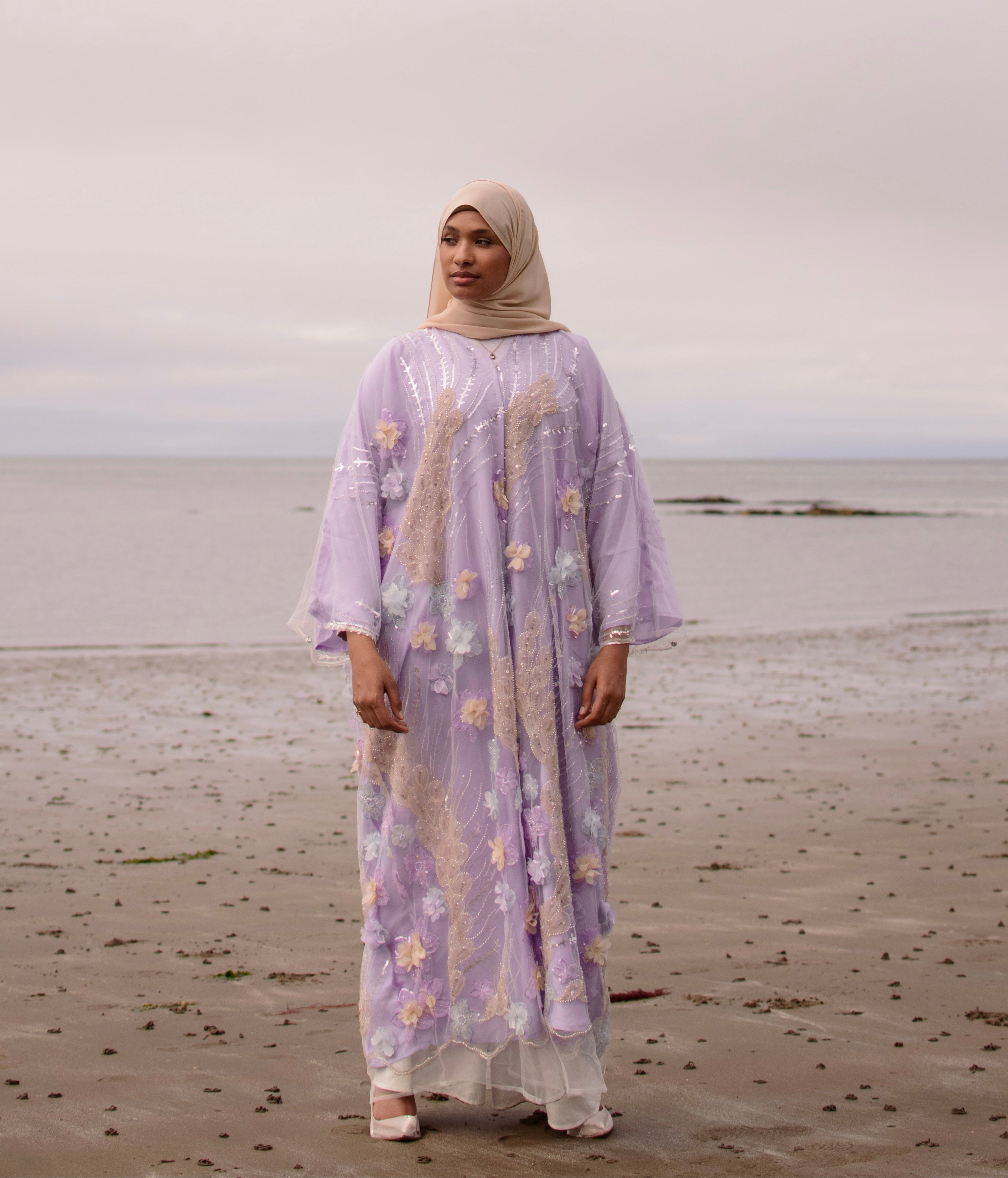 Woman in a long, light-colored dress with floral patterns standing on a beach.