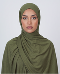 A woman wearing an olive green draped hijab, photographed against a clean light gray studio background.