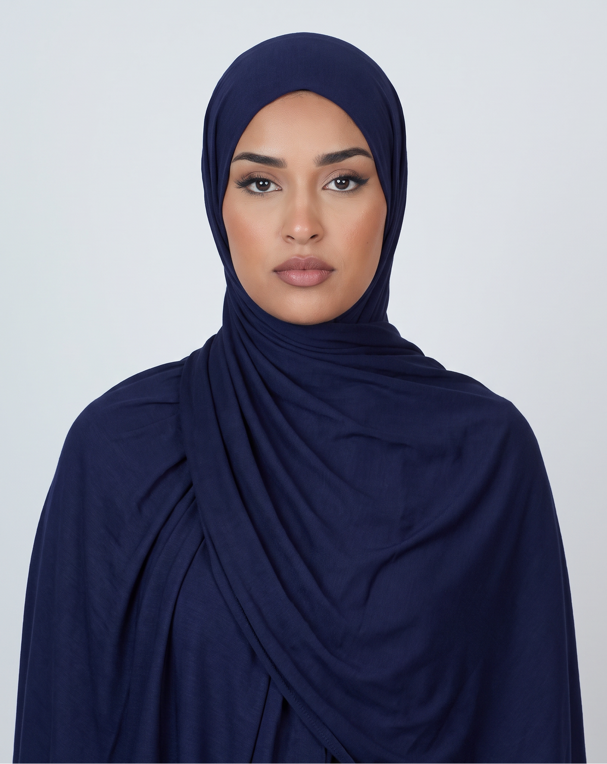 A woman wearing a draped navy blue hijab or shawl, photographed against a clean light gray studio background.
